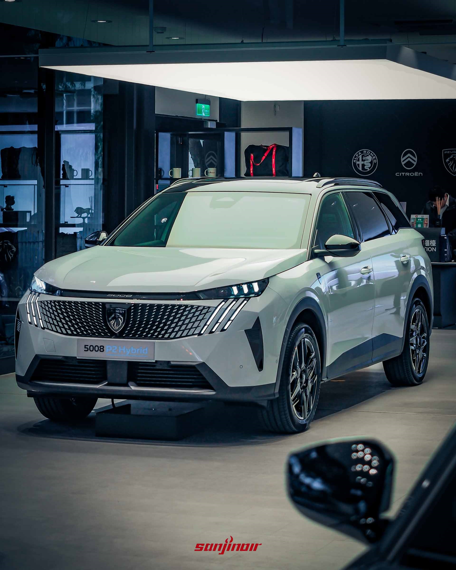 一輛白色的 PEUGEOT 5008 P2 Hybrid SUV。位於發光的燈板下方。車輛具有標誌性的爪痕 LED 大燈，和巨大的車頭格柵。